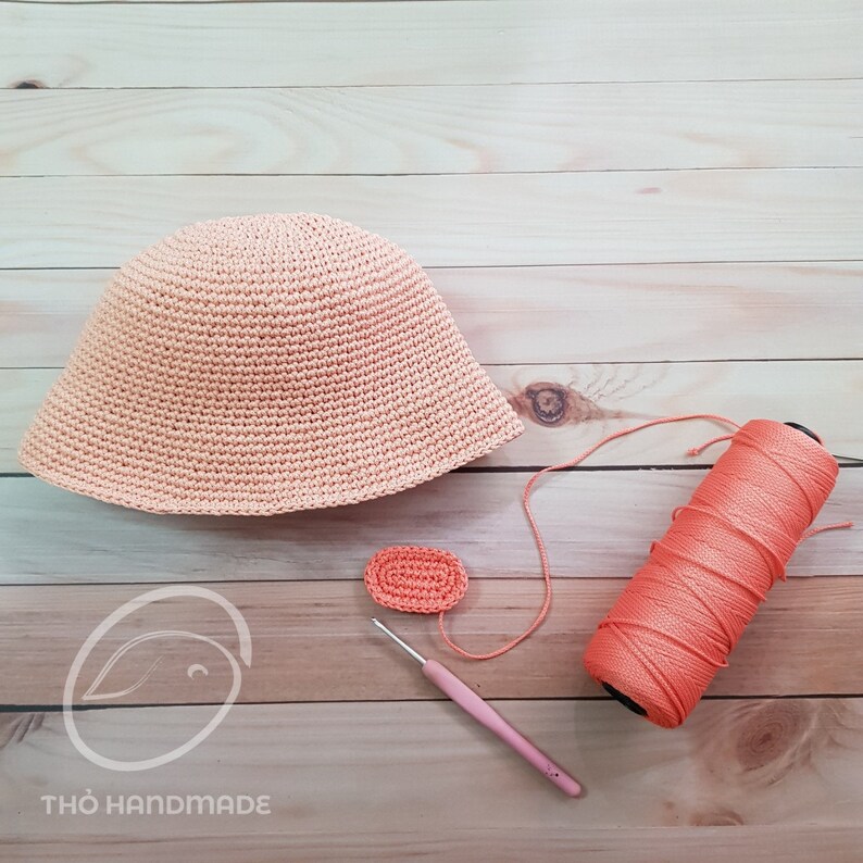 May include: A peach-colored crocheted hat, a small crocheted circle, a ball of peach-colored yarn, and a pink crochet hook on a wooden surface. The text "THƠ HANDMADE" is visible in the bottom left corner of the image.