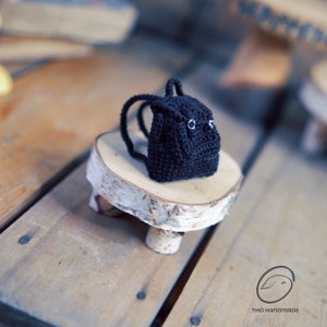 May include: A miniature black crochet backpack with white eye details and a small handle. The backpack is displayed on a small, round wooden stand with three legs. The background is a wooden surface.
