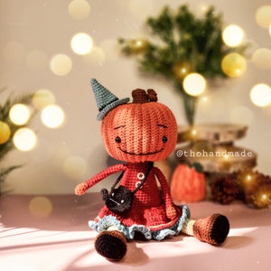 May include: A crocheted pumpkin doll wearing a blue witch hat and a red dress with a black cat purse. The doll is sitting on a pink surface with a blurred background of lights.