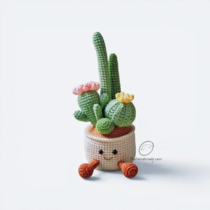 May include: A crocheted cactus plant in a cream-colored pot with a smiling face, two orange feet, and small pink and yellow flowers. The cactus features various shades of green yarn, and the pot displays the text thehandmade.com near the base.