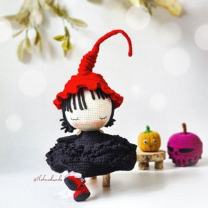 May include: A crocheted witch doll with a red hat, black dress, and red shoes. The doll is sitting on a wooden stool.