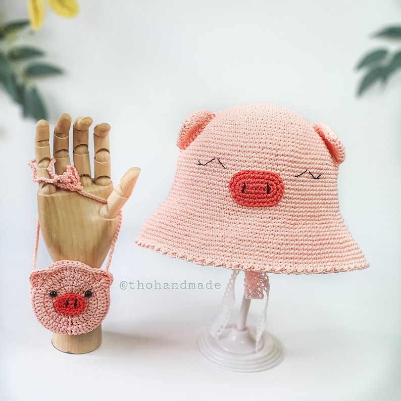 Peut inclure: Un chapeau de cochon en crochet rose avec un petit sac &agrave; main en crochet rose en forme de cochon. Le chapeau a une large bordure et le sac &agrave; main a une bandouli&egrave;re. Le texte "@thohandmade" est visible sur l'image.