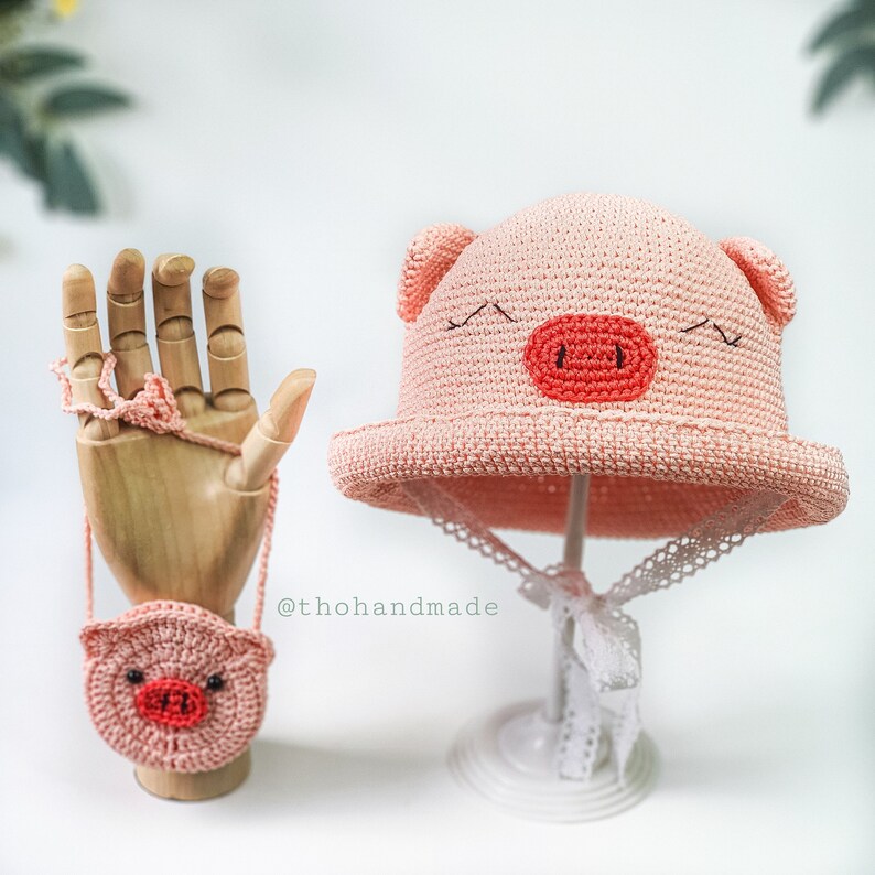 Peut inclure: Un chapeau de cochon rose en crochet avec un petit sac &agrave; main en crochet rose en forme de cochon. Le chapeau est orn&eacute; d'une bordure en dentelle blanche et le sac &agrave; main a une sangle noire.