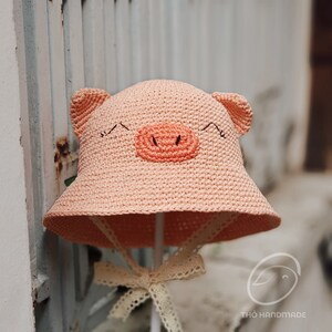 May include: A pink crocheted sun hat with pig ears and a pink pig face. The hat has a white lace trim and a white bow.