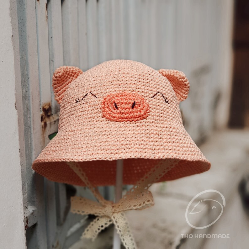 May include: A pink crocheted sun hat with pig ears and a pink pig face. The hat has a white lace trim and a white bow.