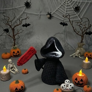 May include: A Halloween-themed display featuring a crocheted Grim Reaper holding a red scythe, surrounded by orange pumpkins, skeletons, and candles. A spider web and bats decorate the background, creating a spooky atmosphere.