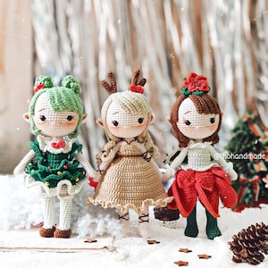 May include: Three crocheted dolls dressed in festive holiday attire. The doll on the left is wearing a green and gold dress with a Christmas tree design. The doll in the middle is wearing a brown dress with a white collar and a red bow. The doll on the right is wearing a red dress with a white collar and a green bow.