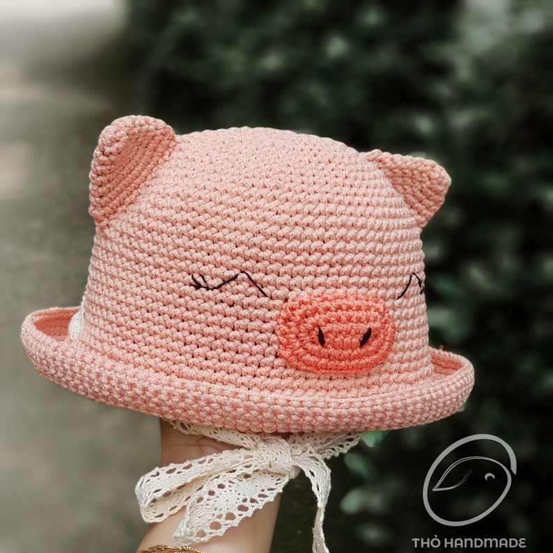 May include: A pink crocheted hat shaped like a pig with black eyes and a pink snout. The hat has a wide brim and a white lace ribbon tied around it.