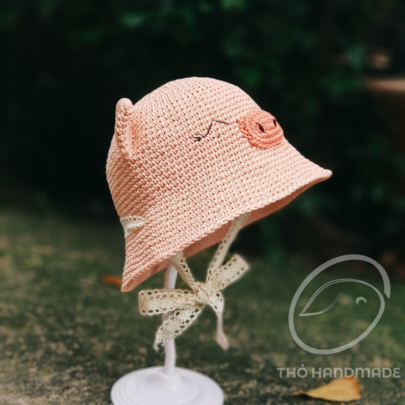 May include: A pink crocheted sun hat with a pig face design. The hat has a white lace trim and ties.