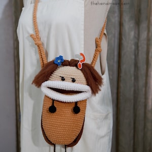May include: A crochet crossbody bag shaped like a character with a tan face, brown hair, and a wide white mouth. It features a blue flower, an orange question mark icon, two black circular buttons, and two dangling black pom-pom feet.