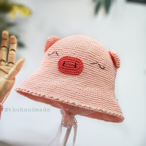 Peut inclure: Un chapeau de soleil en crochet rose avec un motif de visage de cochon. Le chapeau a une large bordure et un petit museau en crochet rose avec des yeux noirs.