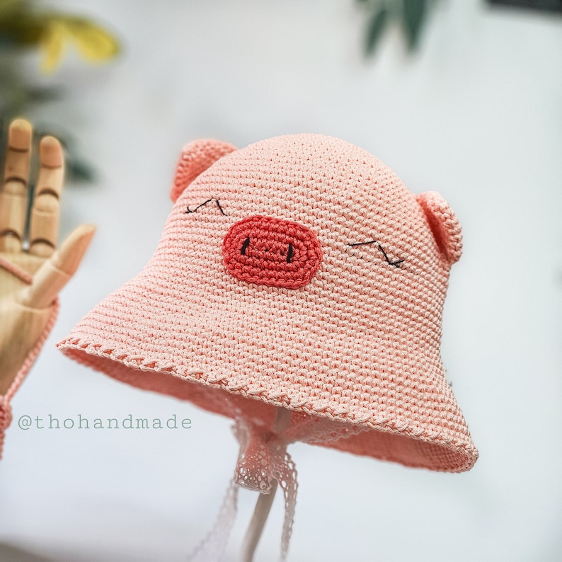 Peut inclure: Un chapeau de soleil en crochet rose avec un motif de visage de cochon. Le chapeau a une large bordure et un petit museau en crochet rose avec des yeux noirs.