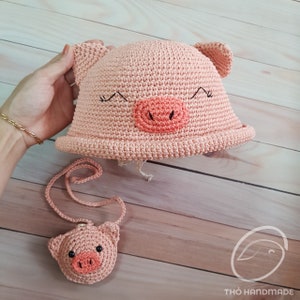 May include: A pink crocheted pig hat with a matching pig keychain. The hat has a wide brim and features embroidered eyes and a pink snout.