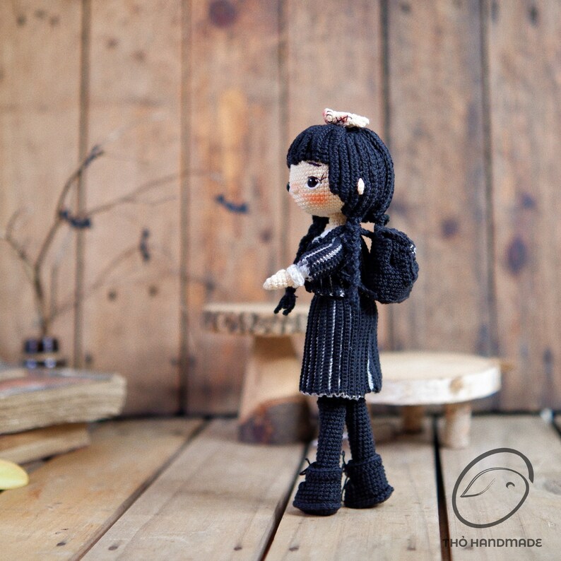 May include: A handmade crochet doll with black hair and a black dress with white stripes. The doll has a small backpack and is standing on a wooden surface. The doll's face is peach-colored with black eyes and a small bow in her hair.