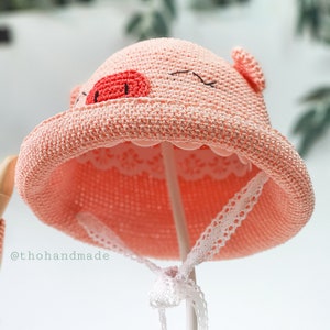 Peut inclure: Un chapeau de soleil en crochet rose avec un motif de visage de cochon. Le chapeau a une bordure en dentelle blanche et est sur un support blanc.