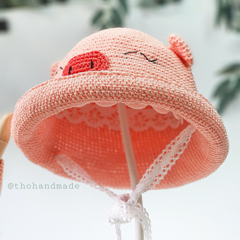 Peut inclure: Un chapeau de soleil en crochet rose avec un motif de visage de cochon. Le chapeau a une bordure en dentelle blanche et est sur un support blanc.