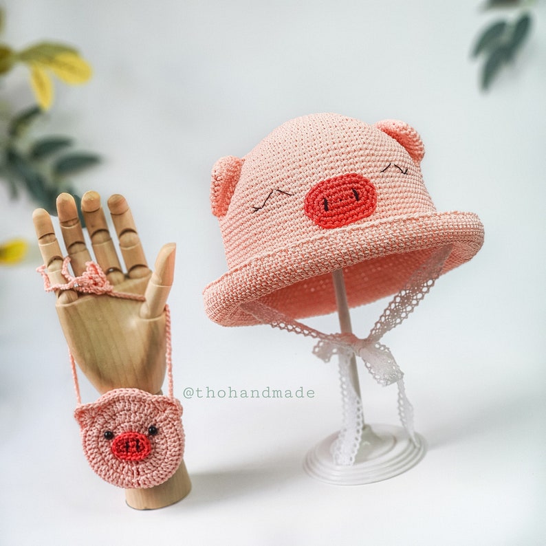 Peut inclure: Chapeau en crochet rose en forme de cochon avec une bordure en dentelle blanche et un sac &agrave; main en crochet rose assorti. Le chapeau est sur un support blanc et le sac &agrave; main est sur une main en bois.