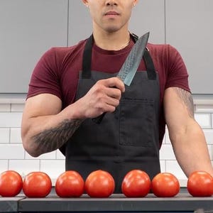Peut inclure: Un homme portant un tablier noir et une chemise rouge tient un couteau de cuisine noir et argent. Devant lui, neuf tomates rouges sont alignées sur une surface sombre.