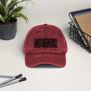 May include: A red vintage style baseball cap with the word "INDIGENOUS" embroidered in black on the front.