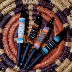 May include: Four black pens with colorful beaded designs. The pens are arranged in a circle on a woven basket.