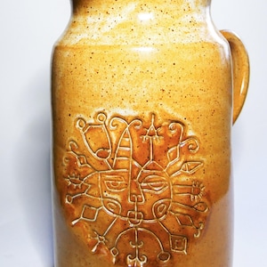Könnte beinhalten: Ein keramischer Krug mit Henkel und Ausguss, verziert mit einem geprägten sonnenartigen Gesichtsdesign. Der Krug ist braun gesprenkelt, mit einem helleren Rand. Das Design ist in einem helleren Braunton gehalten.