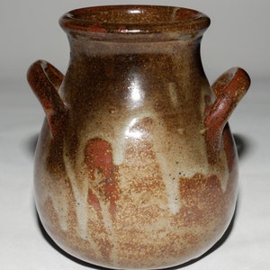 Könnte beinhalten: Eine kleine, braune Keramikvase mit zwei Henkeln. Die Vase hat eine gesprenkelte Glasur mit braunen, weißen und roten Akzenten.