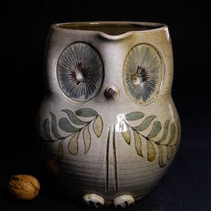 May include: A ceramic owl-shaped pitcher with a cream-colored glaze. The pitcher features large, stylized eyes with blue and brown details, and leaf-like patterns. The owl has a small beak and two vertical lines. A walnut sits to the left.