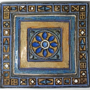 Könnte beinhalten: Quadratische Keramikfliese mit geometrischem Design. Die Fliese zeigt ein zentrales Blumenmotiv in Blau und Weiß, umgeben von einem braunen und blauen Quadrat. Der Rand ist mit kleinen geometrischen Formen in verschiedenen Farben verziert, darunter Blau, Braun und Weiß.
