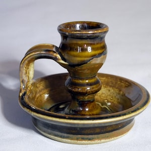 Könnte beinhalten: Ein brauner Kerzenhalter aus Keramik mit einem Griff und einer kleinen Schale. Der Halter hat eine glasierte Oberfläche mit braunen und weißen Streifen.