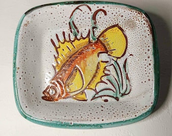May include: A white ceramic dish with a green rim features a hand-painted yellow fish with orange and brown details. The fish is surrounded by green and brown seaweed. The dish has a speckled white glaze.