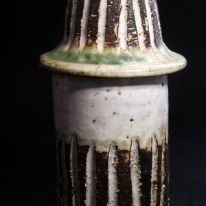 Puede incluir: Un tarro de cerámica artesanal con tapa. El tarro es cilíndrico con ranuras verticales y una combinación de colores blanco y marrón. La tapa tiene forma acampanada con un borde verde. El tarro mide aproximadamente 15 cm de altura.