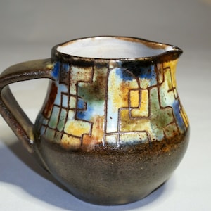 Puede incluir: Un pequeño jarro de cerámica hecho a mano con base y asa de color marrón oscuro. El cuerpo presenta un colorido patrón geométrico en tonos azules, amarillos, verdes y marrones. El interior es blanco y el pico está ligeramente curvado.