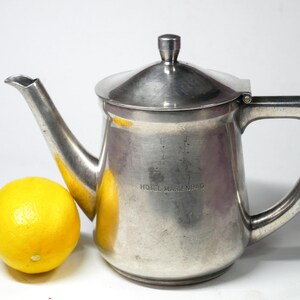 May include: A silver-colored metal teapot with a lid and handle, next to a yellow orange. The teapot has the words "HOTEL MARIENBAD" etched on its side. The teapot has a spout for pouring.