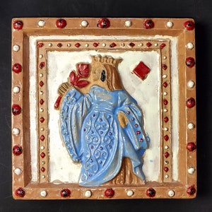 Peut inclure: Un carreau de céramique avec une image en relief d'une reine tenant une fleur. La reine porte une robe bleue et blanche avec une couronne. Le carreau est entouré d'un cadre brun avec des points rouges et blancs.