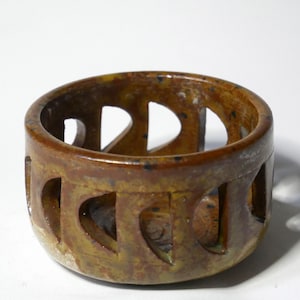 Könnte beinhalten: Ein runder Kerzenhalter aus Holz in Braun- und Gelbtönen. Der Halter hat herzförmige Aussparungen an den Seiten. Die Holzmaserung ist sichtbar und verleiht ihm ein rustikales Aussehen. Geeignet für eine kleine Kerze.