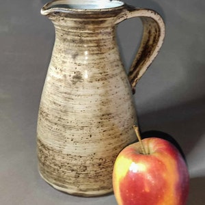 Pode incluir: Um jarro de cerâmica marrom e branco com uma boca larga e uma alça. O jarro tem um esmalte salpicado e está sentado em uma superfície cinza ao lado de uma maçã vermelha.