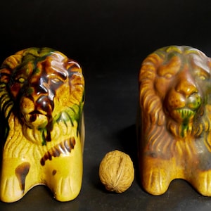 Könnte beinhalten: Zwei Löwenfiguren aus Keramik mit gelber, brauner und grüner Glasur. Die Löwen haben detaillierte Mähnen und sind aufrecht positioniert. Zwischen den Löwen liegt eine Walnuss. Die Figuren sind dekorativ und wahrscheinlich vintage.