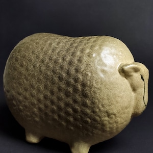 May include: A ceramic sheep figurine with a textured, speckled surface in shades of brown and beige. The sheep has a rounded body and four short legs.