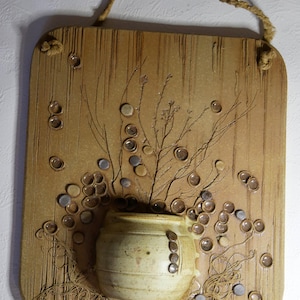 Könnte beinhalten: Ein Keramik-Wandbild mit braunem Hintergrund und einem kleinen weißen Topf mit braunem Rand. Der Topf ist umgeben von braunen und grauen Kreisen und einer getrockneten Pflanze mit braunen Stielen.