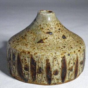 May include: A small, ceramic vase with a wide base and a narrow neck. The vase is decorated with a brown and white geometric pattern and has a speckled glaze.