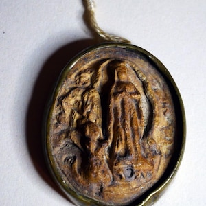 Puede incluir: Un colgante ovalado de cerámica marrón con una imagen en relieve de una mujer con una túnica larga. El colgante está rodeado por un marco de metal y tiene una cuerda atada a la parte superior.