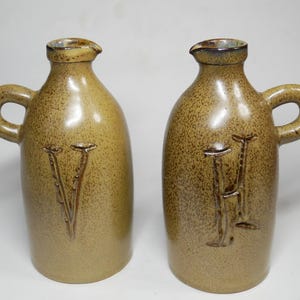Puede incluir: Dos jarras de cerámica con esmalte beige moteado, cada una con un asa lateral y un pequeño pico vertedor. Una jarra tiene una letra V en relieve en la parte frontal, mientras que la otra jarra presenta una letra H en relieve en la superficie.