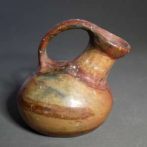 Puede incluir: Una pequeña jarra de cerámica marrón con un cuerpo ancho y redondeado y un cuello corto y estrecho. La jarra tiene un asa que se curva hacia arriba y sobre el cuello. La jarra está decorada con rayas de pintura roja, marrón y blanca.