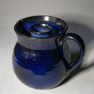 Könnte beinhalten: Ein blauer Kerzenhalter aus Keramik mit Henkel und einer runden Öffnung für eine Kerze. Der Halter hat eine gesprenkelte Glasur.