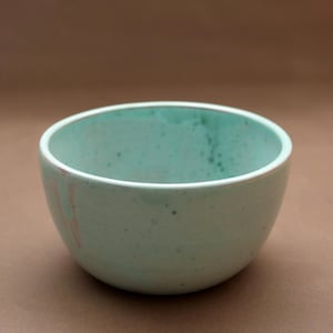 May include: A round ceramic bowl with a light mint green glaze featuring subtle dark speckles throughout the surface. The interior and exterior display a smooth finish with a faint pinkish streak visible on the left side of the curved wall.