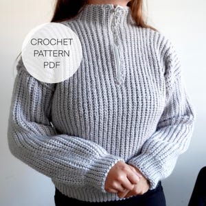 May include: A light gray, ribbed knit sweater with a high collar and a silver zipper. The sweater has long sleeves and a cropped fit. The text "CROCHET PATTERN PDF" is in a white circle on the left side of the sweater.