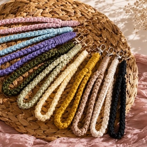 Può includere: Una collezione di cordini da polso in macramè colorati in varie tonalità, tra cui rosa, blu, viola, verde, giallo, marrone, bianco e nero. Ogni cordino presenta un design intrecciato e una chiusura color argento. I cordini sono disposti su un cestino intrecciato.