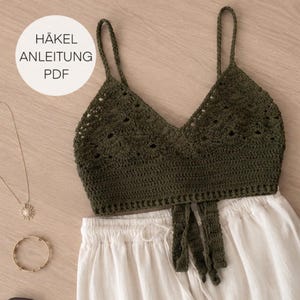 Op de afbeelding: Een olijfgroene gehaakte crop top met dunne bandjes, een V-hals en een schulprandpatroon, voorzien van twee bungelende linten aan de voorzijde. Een witte cirkelvormige afbeelding met de tekst HÄKEL ANLEITUNG PDF staat in de linkerbovenhoek.