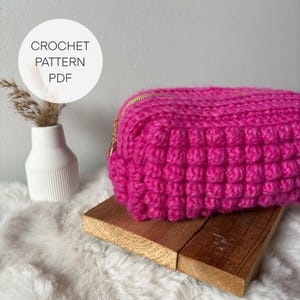 May include: A bright pink crocheted pouch with a gold zipper. The pouch is sitting on a wooden surface.