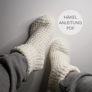 May include: A pair of white crocheted slipper socks. The socks are made with a fluffy yarn and have a textured pattern. The image includes the text "HÄKEL ANLEITUNG PDF".